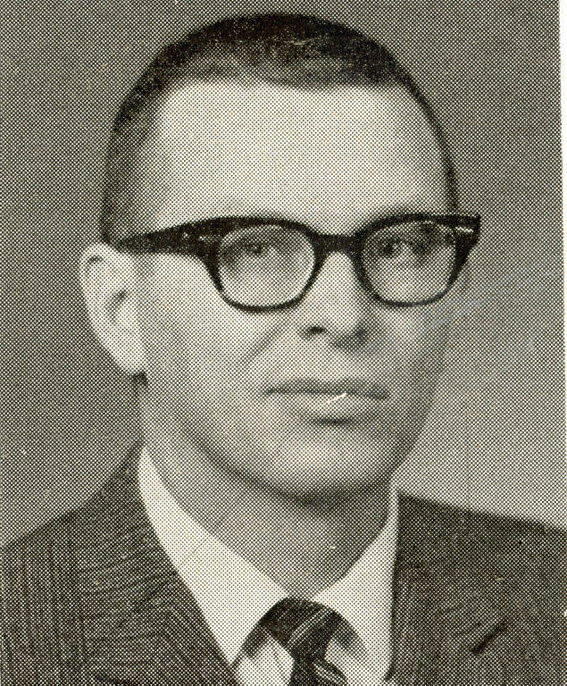 A man with short hair and glasses, wearing a suit and tie, is facing forward with a neutral expression.