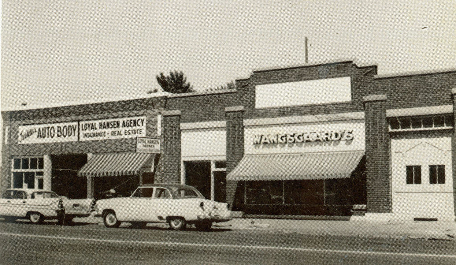 A street view of three adjacent brick buildings. The first building has a sign that reads "Loyal Hansen Agency Insurance - Real Estate" and "Auto Body," with a striped awning. Two cars are parked in front. The second building has a large sign with "Wangsgaard's" and a striped awning. The third building has a plain façade with no visible signage. Trees are partially visible in the background.