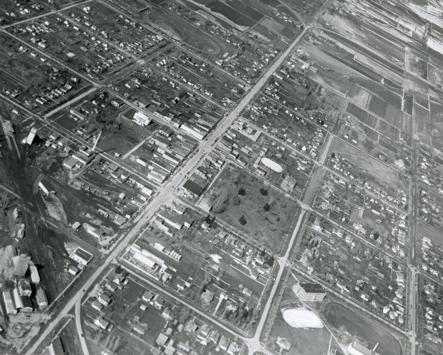 An aerial view of a city layout showing a grid of streets with various small buildings and houses. There are patches of open land and fields. The intersections are clearly visible, and there are some larger buildings interspersed among smaller structures.