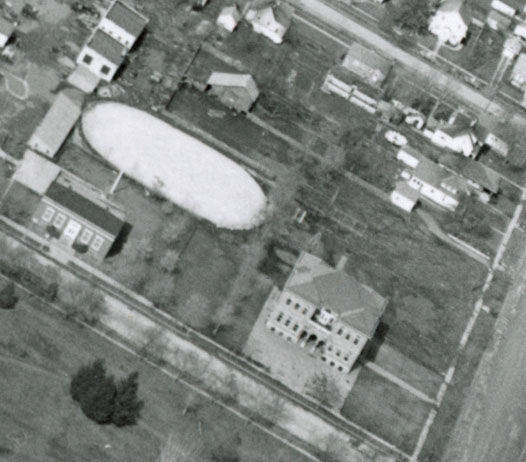 Aerial view of a neighborhood with several buildings and houses. There is a distinct oval-shaped area that appears to be a pond or field. Roads and a grid layout of the area are visible, with some trees and greenery scattered throughout. One prominent building with a decorative facade is positioned near the center right.