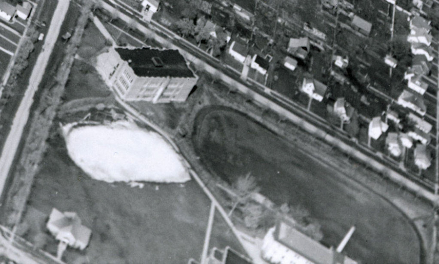 An aerial view of a landscape featuring a large rectangular building and an adjacent oval-shaped area. To the right, a series of small residential houses are lined along a street. There are various roads and patches of grassy areas surrounding the structures.