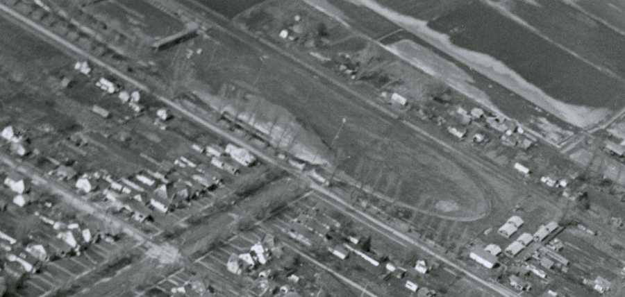 An aerial view of a neighborhood with rows of small houses lined along streets. There is an oval-shaped area that appears to be a track or park surrounded by more houses. Fields can be seen in the background.