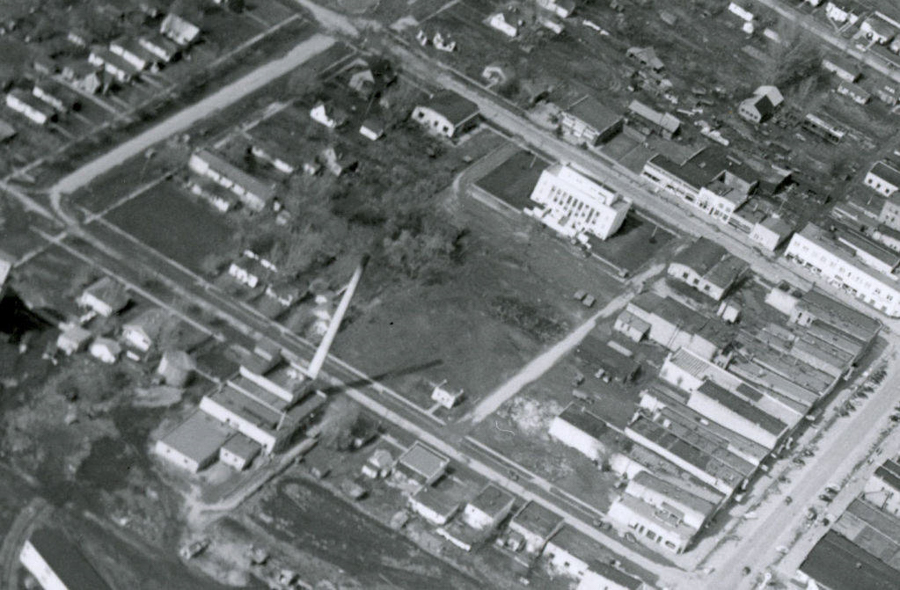 An aerial view of a town featuring grid-like streets lined with small buildings. A larger, prominent building with a front staircase is situated within a spacious yard area. Roads and intersections divide residential areas, with scattered houses and open plots visible. There are several small structures and trees dispersed throughout the scene.