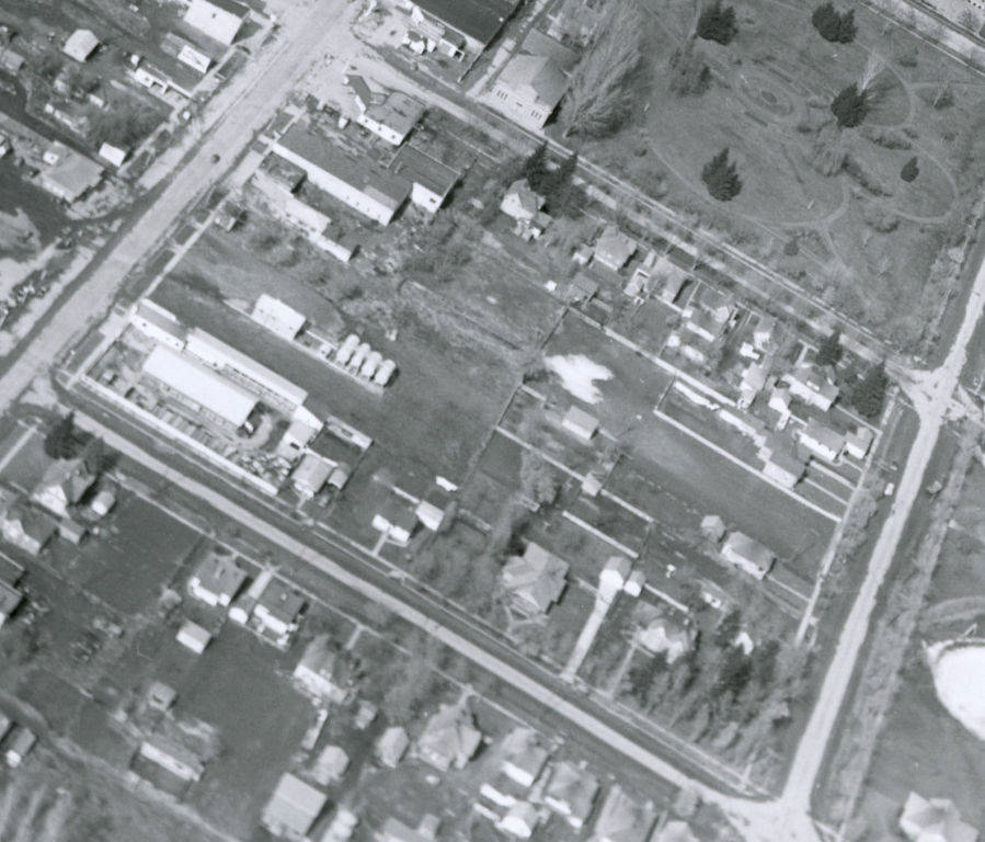 An aerial view of a neighborhood with intersecting streets and rows of small buildings and houses. There is a large open area with pathways and scattered trees, which appears to be a park, occupying part of the scene. The layout shows a mixture of residential and possibly industrial or commercial structures.