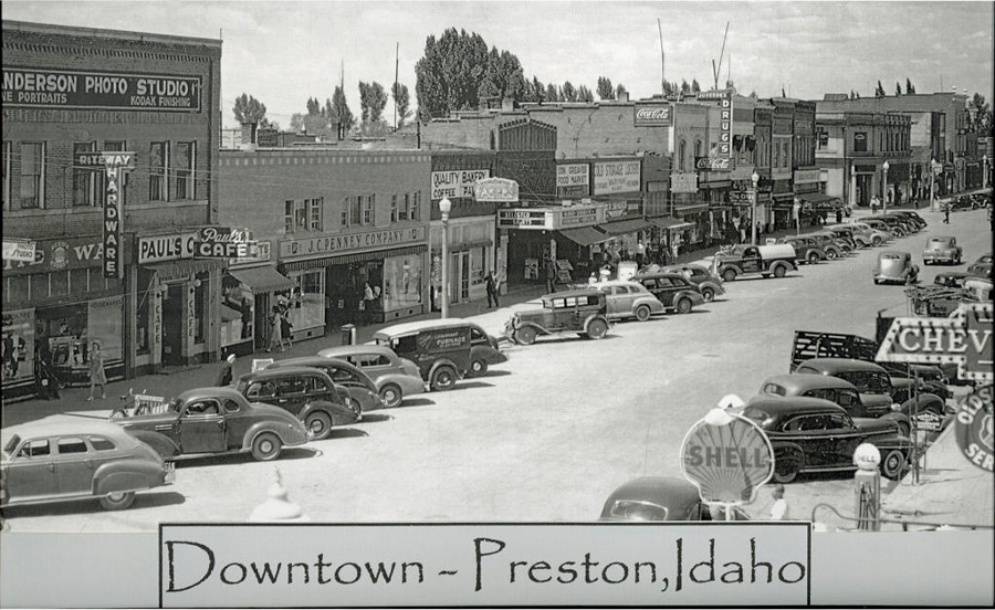 A bustling street scene in downtown Preston, Idaho, featuring vintage cars parked along both sides of the road. The street is lined with various businesses, including a photo studio, hardware store, Paul's Cafe, J.C. Penney Co., a drug store, and a bakery. Several people are walking on the sidewalk. A Shell gas station sign is prominently visible.