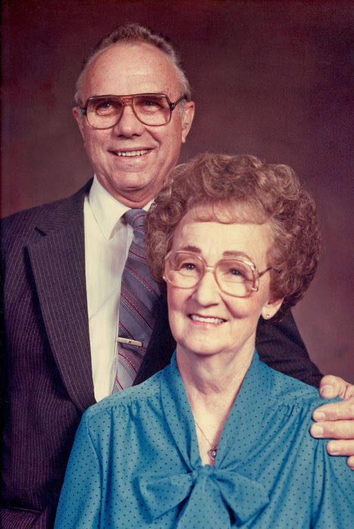 An older man and woman are posing together. The man is standing slightly behind the woman with his hand on her shoulder, wearing a suit and tie with glasses. The woman is in front, also wearing glasses and a blouse with a bow at the neck. They are both smiling.