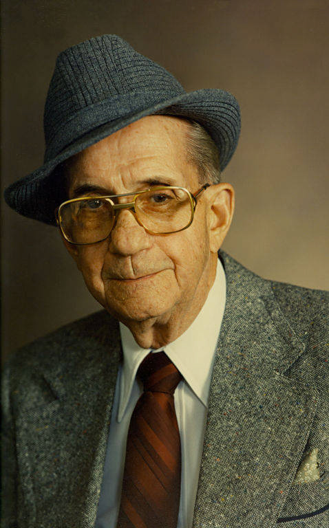 An elderly man wearing a gray suit and tie, a white shirt, a gray fedora, and large glasses.