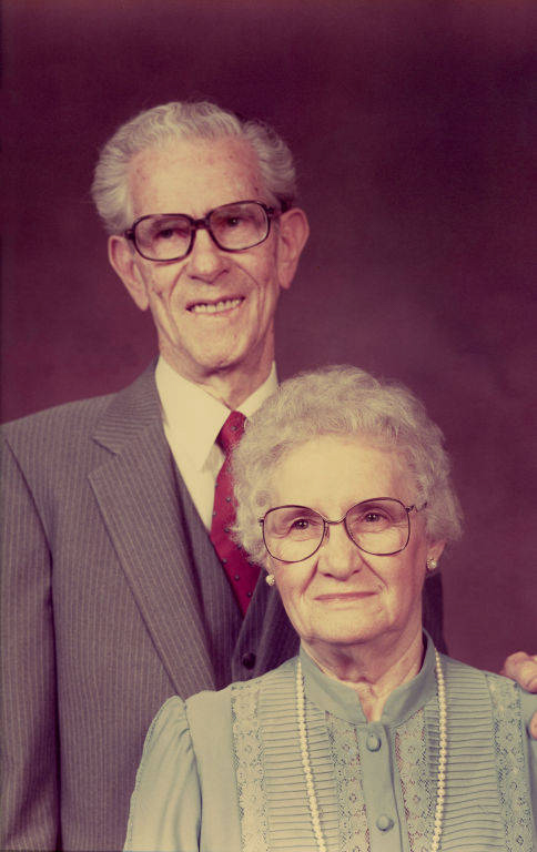 An elderly couple standing together. The man is wearing a suit, shirt, and tie, and has glasses. The woman is wearing a blouse with lace detailing, a necklace, and glasses. Both have white hair and are looking at the camera.