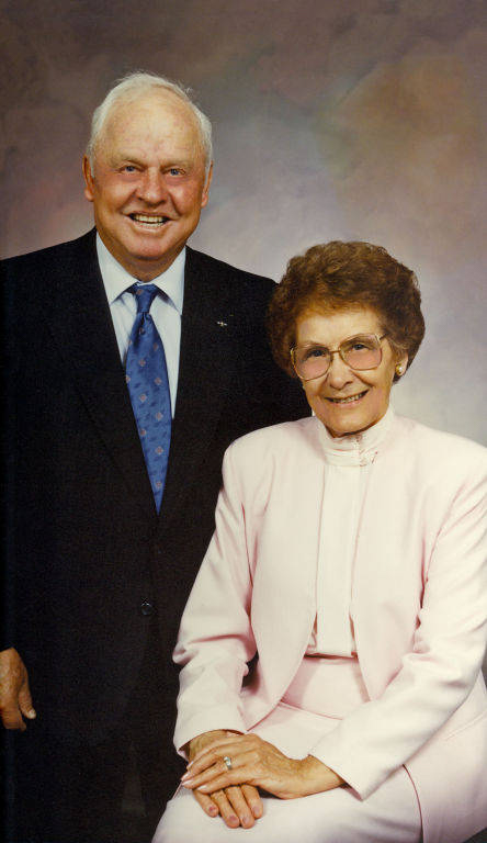 An older man and woman are posing together. The man is standing and wearing a dark suit with a blue tie, while the woman is seated, dressed in a light pink outfit, and wearing glasses. They are both smiling at the camera.