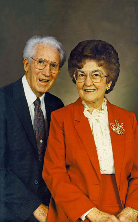 An elderly man and woman are posed together. The man is wearing a dark suit with a patterned tie and glasses. The woman is wearing a red suit with a white blouse and glasses, and she has a decorative pin on her jacket. Both are smiling.