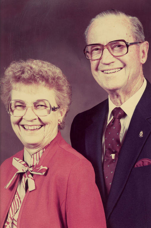 An elderly couple is standing side by side, both wearing glasses and smiling. The woman is wearing a red jacket with a striped bow around her neck, and the man is dressed in a dark suit with a tie and a small pin on his lapel.