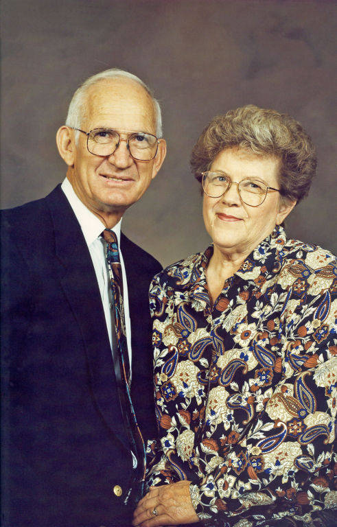 An older couple standing close together and smiling. The man is wearing a dark suit, white shirt, and patterned tie, with glasses. The woman is wearing a patterned blouse and glasses, with her hair styled short.