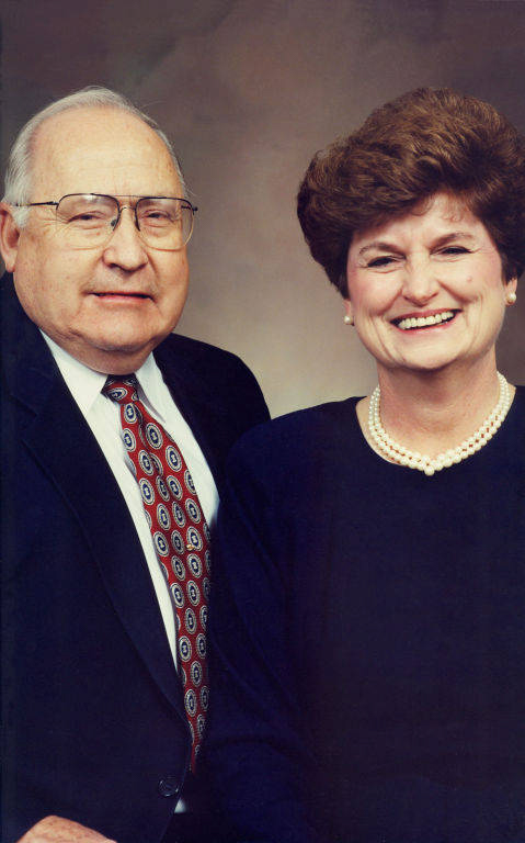 An older man and woman smiling and standing side by side. The man is wearing glasses, a dark suit, a white shirt, and a patterned tie. The woman is wearing a dark top with a pearl necklace and earrings.