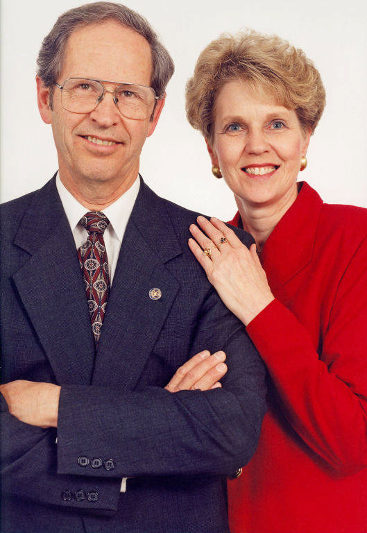 A man in a suit with a patterned tie, standing with arms crossed. A woman stands next to him, smiling, with one hand on his shoulder. She is wearing a red outfit. Both are looking at the viewer.