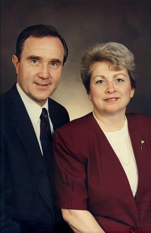 A man and a woman are posing together. The man is wearing a dark suit and tie, and the woman is wearing a red blazer with a pearl necklace. They both have light-colored hair and are looking forward.
