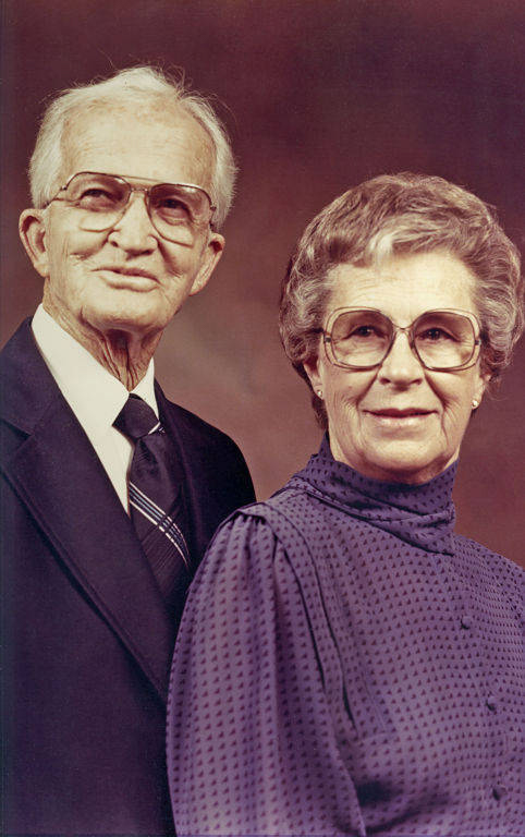 An elderly man and woman standing closely side by side, both wearing glasses. The man is dressed in a suit with a tie, while the woman is wearing a patterned blouse with a high collar. They are both looking toward the camera.