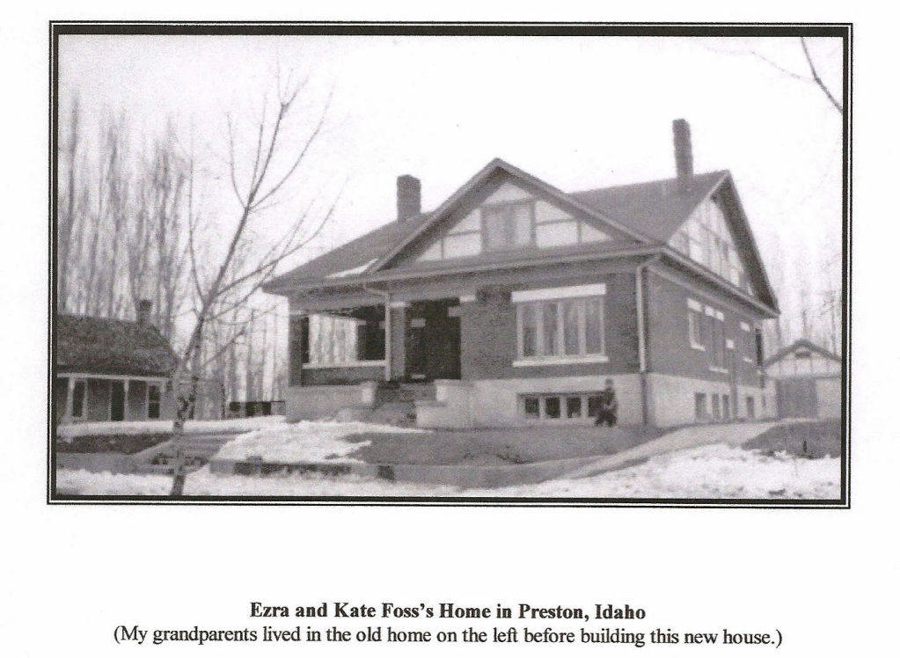 A large two-story house with a porch and multiple windows is situated on a snowy landscape. There is a smaller building to the left of the main house. A person is sitting on the steps in front of the house. Sparse trees are visible in the background. Text reads: "Ezra and Kate Foss's Home in Preston, Idaho." Below it, another line reads: "(My grandparents lived in the old home on the left before building this new house.)"