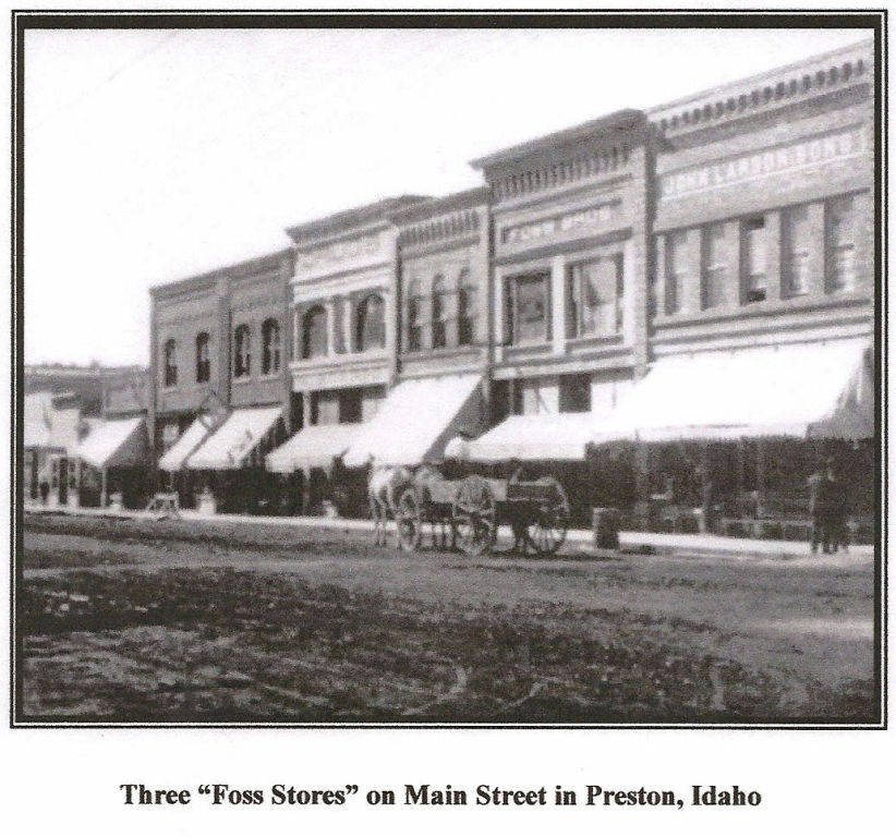 A row of historic-style buildings with awnings along a dirt street. There is a horse-drawn wagon in front of the buildings. Two signs on the buildings read "Foss Hardware & Sons" and "R. R. Green & Sons." Below the image, the text reads, "Three 'Foss Stores' on Main Street in Preston, Idaho."