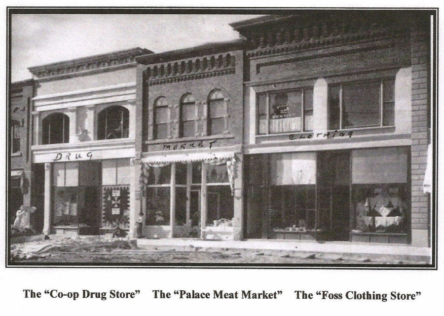 Three storefronts are visible in a row. The first is labeled "DRUG," the second is labeled "MARKET," and the third is labeled "CLOTHING." Below the image, there are captions for each store, reading: "The 'Co-op Drug Store,'" "The 'Palace Meat Market,'" and "The 'Foss Clothing Store.'"