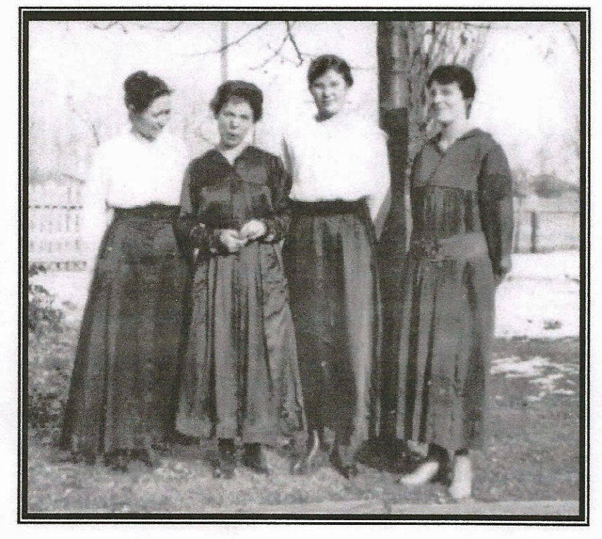 Four women standing in a garden, wearing long skirts and blouses. They are posed side by side with trees and a fence in the background.