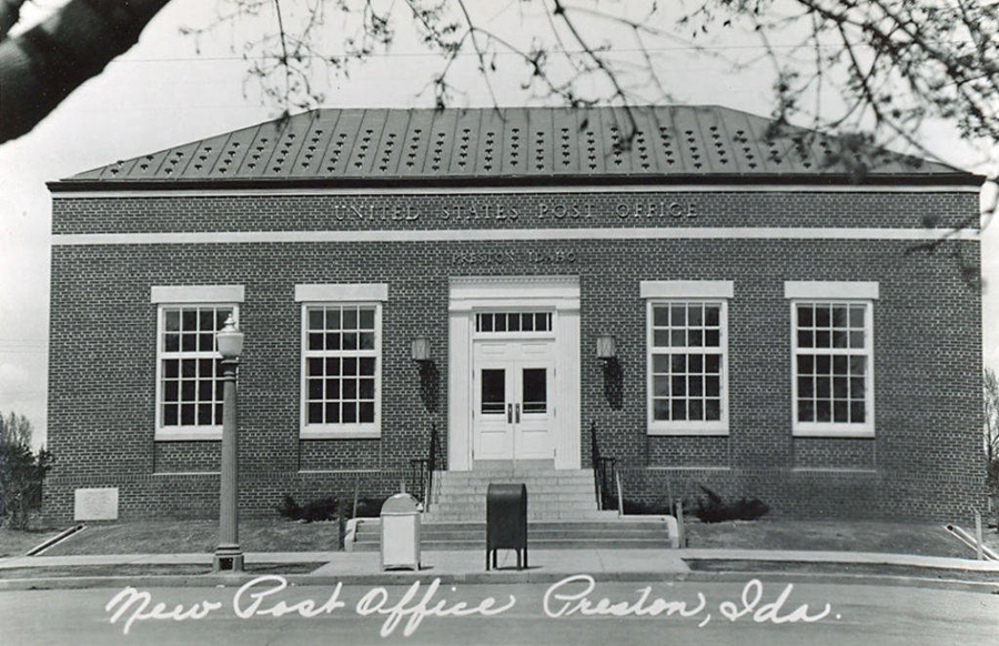 A brick building with a sign reading "United States Post Office" and "Preston, Idaho." It features large windows symmetrically placed on either side of the central entrance. There are stairs leading up to the entrance with a handrail. The scene is framed by tree branches, and text at the bottom reads "New Post Office, Preston, Ida."