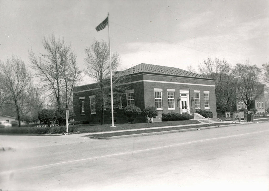 A brick building with the words "United States Post Office" above the entrance. Steps lead up to the door, and windows line the walls. There's a flagpole with an American flag in front. Trees and shrubs are around the building, and a mailbox is visible nearby.