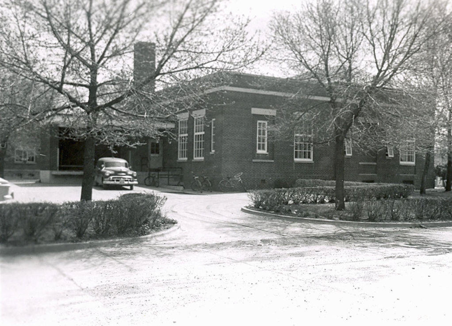 A brick building with large windows and a chimney, surrounded by leafless trees and shrubs. A vintage car is parked near the entrance, and a bicycle is leaning against a railing. The driveway curves around the front of the building.