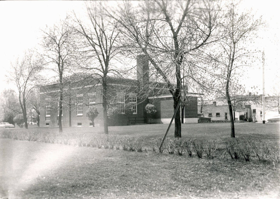 A brick building with large windows surrounded by leafless trees and a grassy lawn. There are bushes lined up in front of the building. A chimney is visible on the structure, and there are a few cars parked in the background.