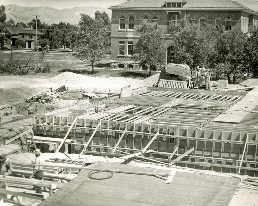 A construction site with wooden framework in the foreground and two workers. A truck and construction equipment are nearby. In the background, there is a large brick building with arched windows, and trees nearby. Houses and mountains can be seen in the distance.
