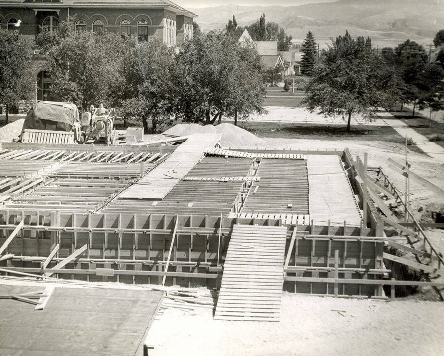A construction site with wooden framework and ramps. In the background, several trees and a large brick building with arched windows are visible. There is a street lined with trees and residential houses further back. A small work vehicle is parked near a pile of materials.