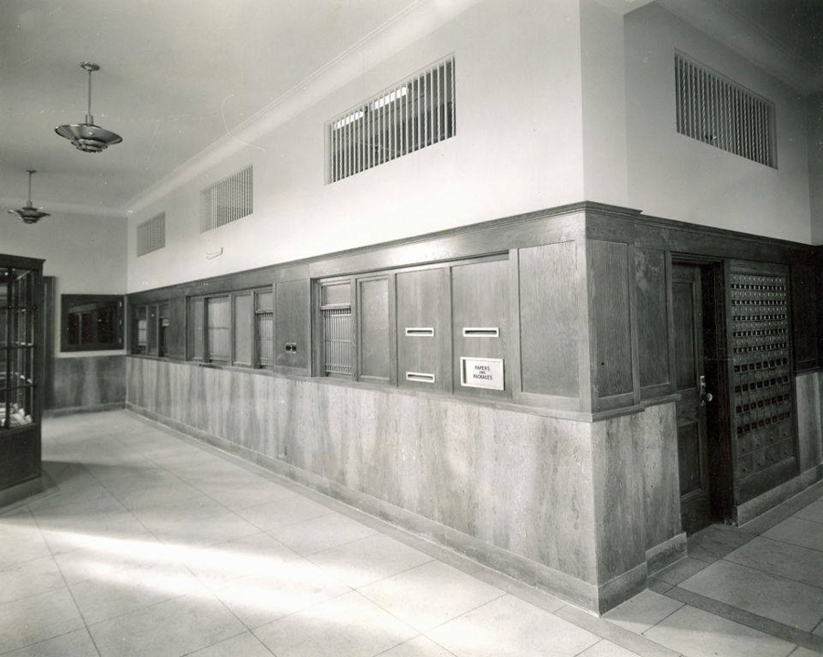 A hallway with a tiled floor and a wood-paneled wall. There are several mail slots and a sign on the wall that reads "PAPERS FILLED IN CUBBIES." A wooden door with small rectangular sections is on the right. Ceiling lights are visible above.