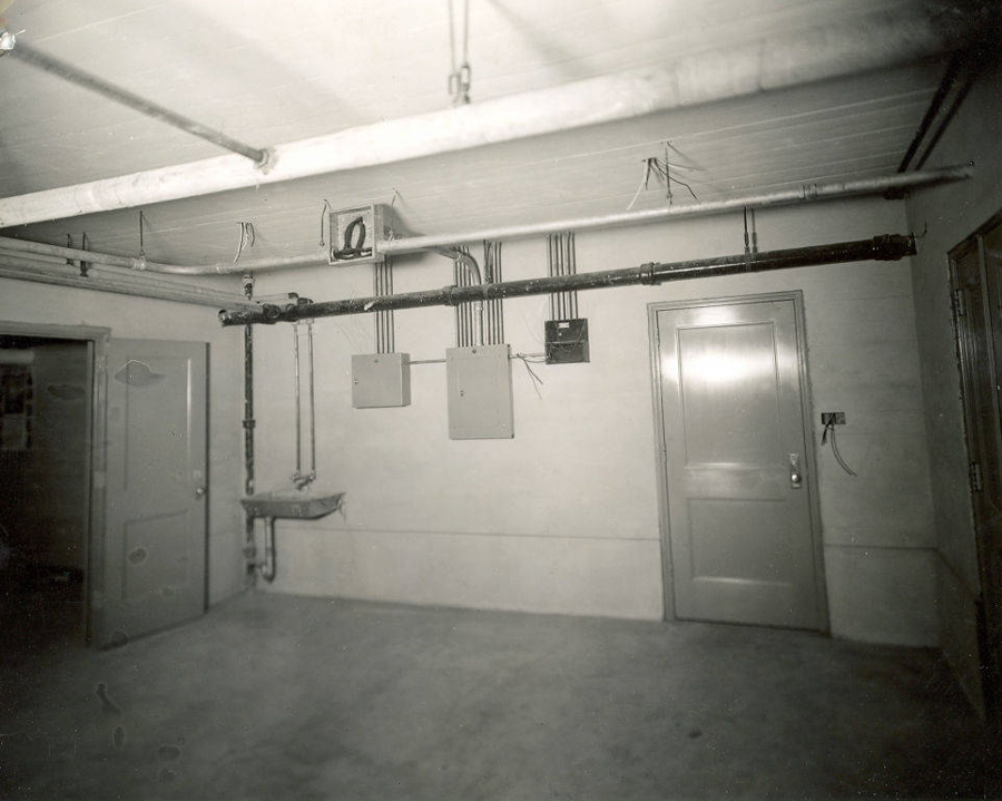 A room with a concrete floor and unfinished walls, featuring two closed wooden doors and one open door. Several pipes and electrical boxes are mounted on the walls. A small sink is attached near the open door, and exposed wiring and pipes run across the ceiling. A light switch with exposed wiring is on the wall near the closed door.