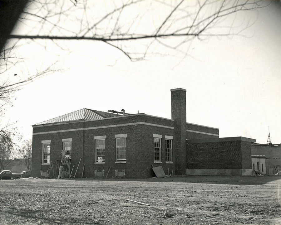 A single-story brick building with large windows and a flat roof. There is a tall chimney on one side. Several people are positioned on scaffolding working near the windows. The foreground is a barren dirt area, and some cars are visible in the background. Bare tree branches overhang the scene.
