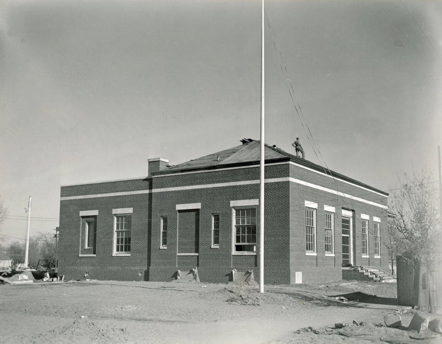 A brick building with large windows and steps leading to an entrance. A person is visible standing on the roof. The surrounding area appears to be a construction site, with dirt and scattered materials. A flagpole is in the foreground.