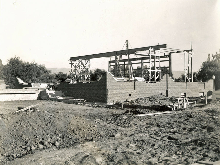 A partially constructed brick building with steel framing and wooden supports. Surrounding the structure are piles of dirt and construction materials. Trees and shrubs are visible in the background.