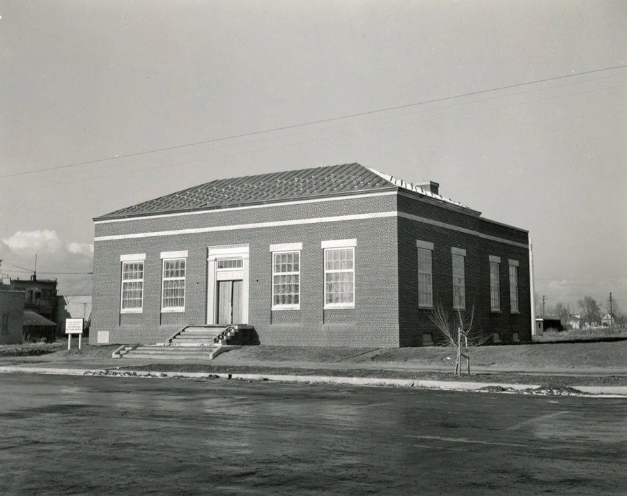 A rectangular brick building with multiple large windows and a central entrance featuring steps leading up to it. There are two signs visible on the left side of the building. The structure sits on a grassy area with a paved street in front. Some other buildings and utility poles are visible in the background.
