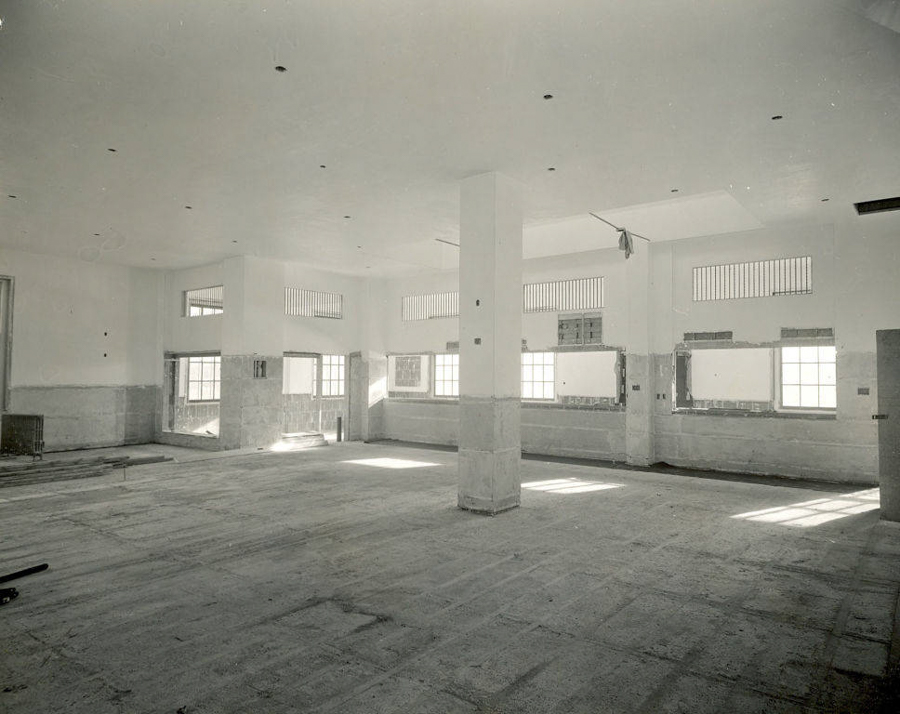 An empty interior space with high ceilings and several large windows. The floor appears unfinished and there are exposed square pillars throughout the room. There are small rectangular windows near the ceiling and a door on one wall. Partially constructed walls are visible, and scattered materials lie on the ground.