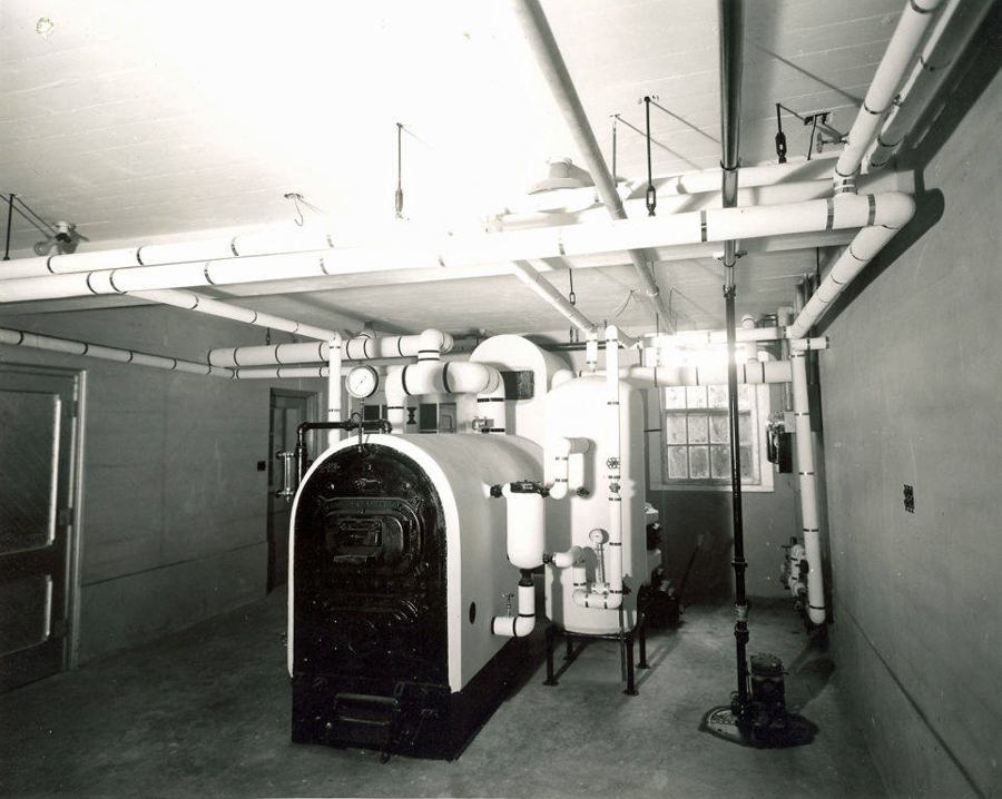 A room containing a large industrial boiler surrounded by numerous pipes running along the ceiling and walls. A pressure gauge is visible on the machinery. In the background, a window allows light into the space. The walls and floor are made of concrete.