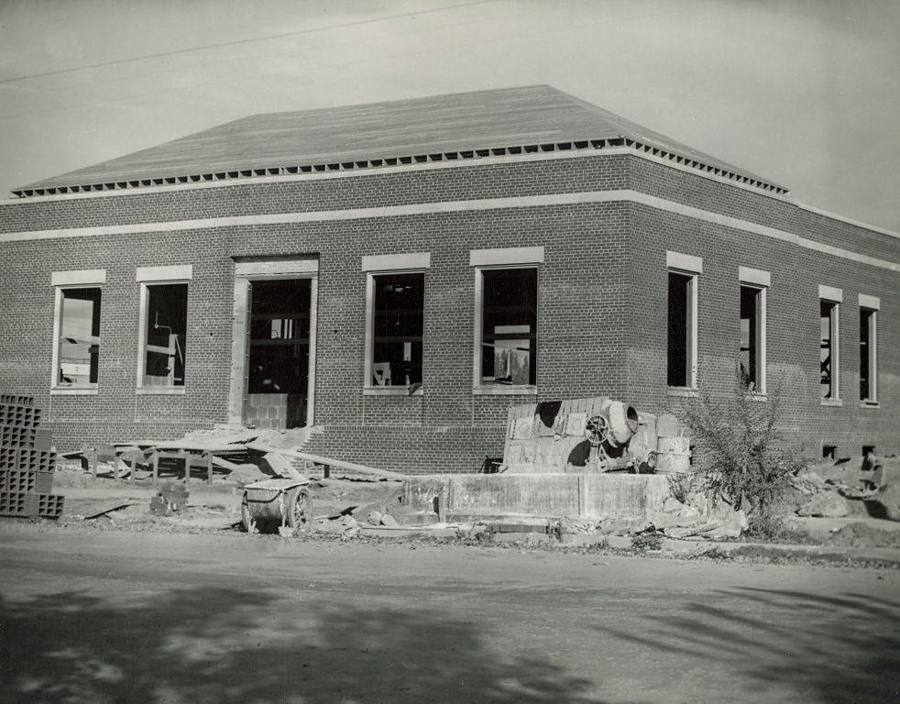 A single-story brick building under construction with several large windows and an open entrance. Construction materials and equipment are scattered around the site, including a cement mixer and stacks of bricks. The ground surrounding the building is unpaved, and there are no trees or landscaping.