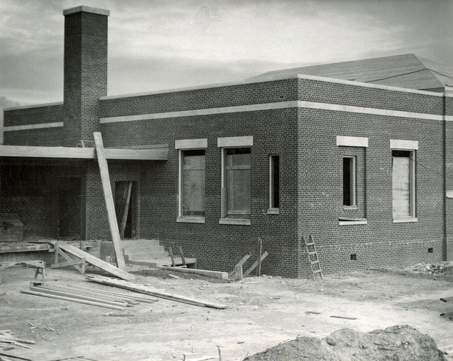 A brick building under construction with several boarded-up windows and a chimney. There is unfinished ground in the foreground with construction materials scattered, including wooden planks and a ladder leaning against the building.