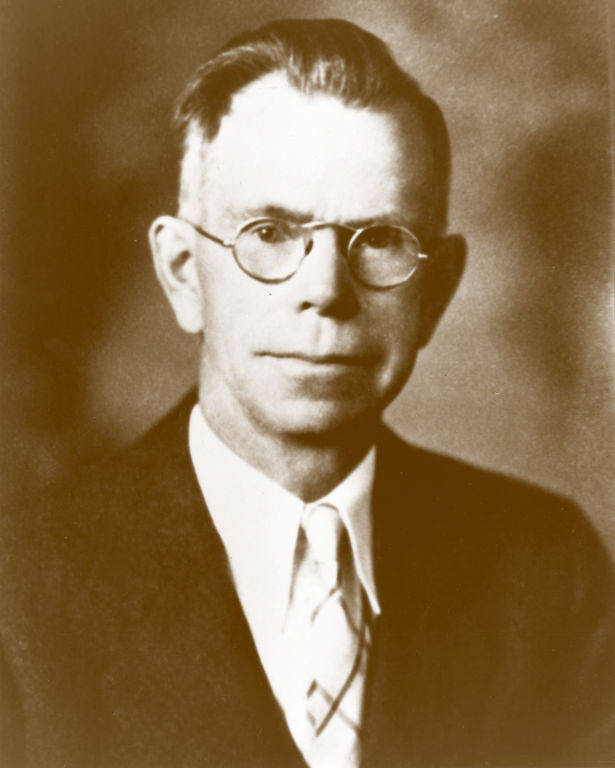 An older man wearing round glasses and a suit with a tie. His hair is neatly combed, and he has a serious expression. There is no text visible in the image.
