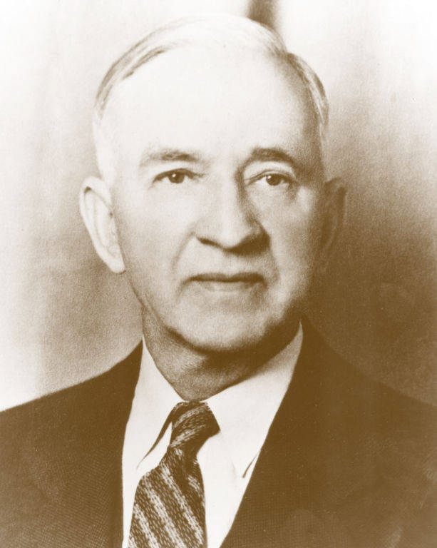 An older man with short hair, wearing a suit and tie, is looking forward.