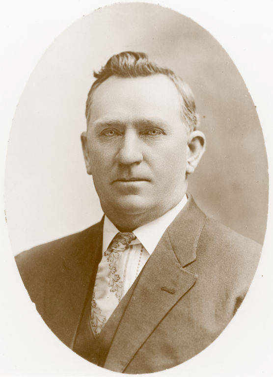 A man wearing a suit and tie with a patterned design, looking directly at the viewer. He has short hair and a solemn expression. The background is plain.