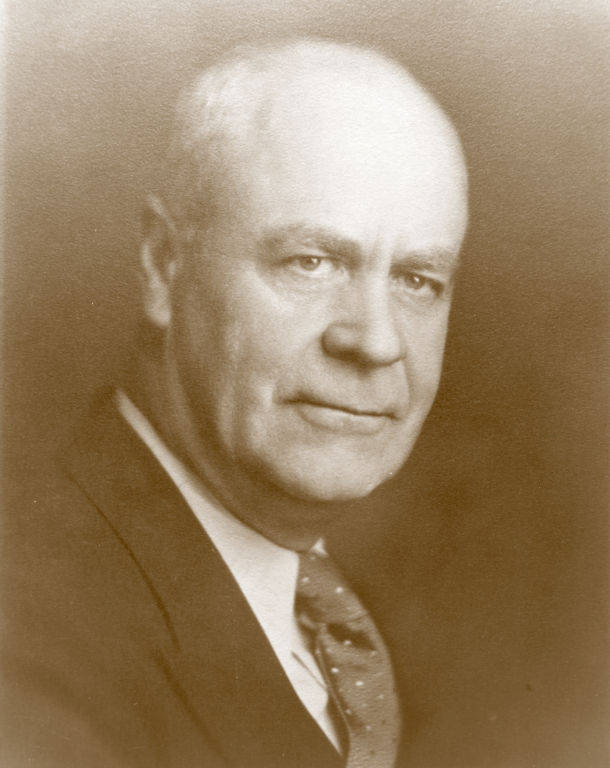 A man dressed in a suit and tie, looking towards the camera with a neutral expression.