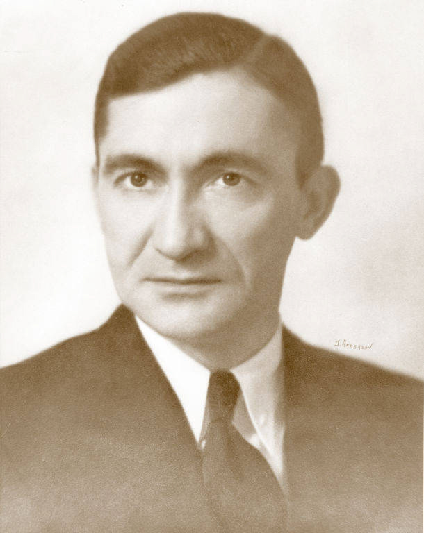 A man wearing a suit and tie is looking directly at the viewer. His hair is combed back, and he has a serious expression. There is a signature in the bottom right corner.