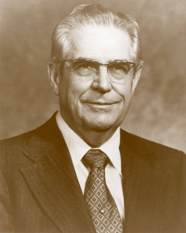An older man with glasses is wearing a dark suit, a white collared shirt, and a patterned tie. He has short, neatly combed gray hair and is smiling slightly. The background is plain.