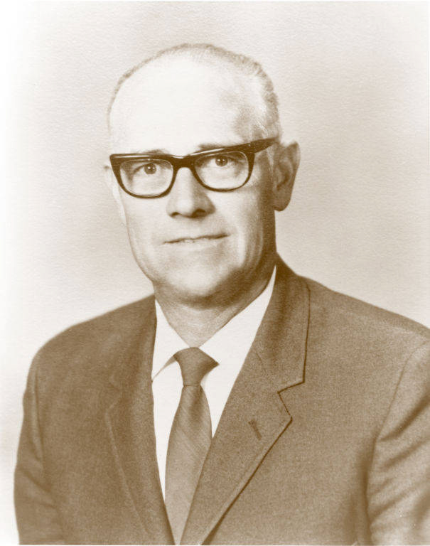 An older man wearing glasses, a suit, white shirt, and a tie, looking straight ahead with a neutral expression.