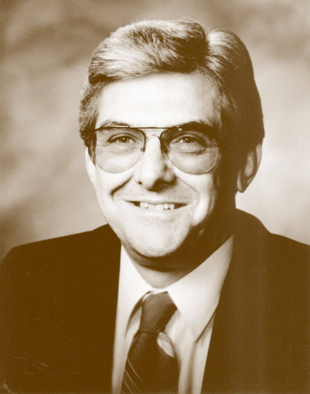A man with short hair and glasses is wearing a suit and tie, smiling.