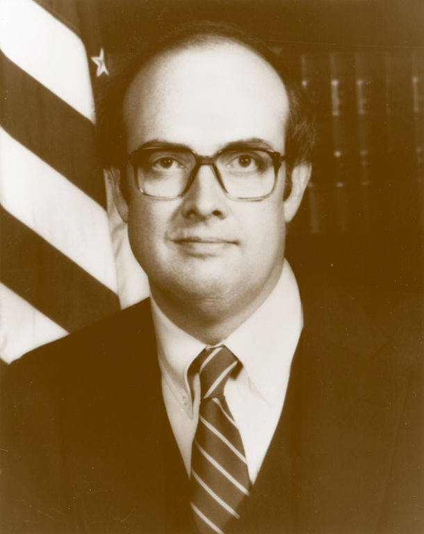 A man wearing glasses and a suit with a striped tie stands in front of a U.S. flag and a background of bookshelves.