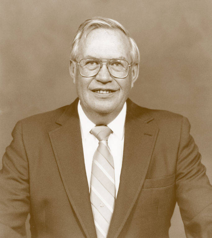An older man wearing glasses, a suit, and a striped tie is smiling and looking towards the camera. He's standing or sitting against a plain background.